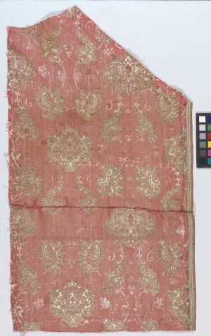  An image of a rectangular piece of dusty-rose textile with an uneven angular cut on the upper right side, adorned with symmetrical, metallic gold floral and vegetal motifs arranged in horizontal bands, accompanied by a color scale indicator to the side.