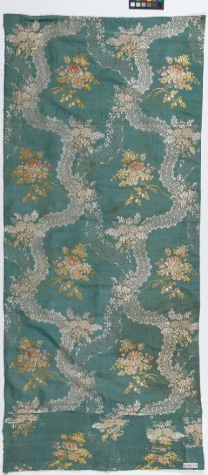  An antique textile with intricate patterns in teal, soft browns, and pale greens, featuring vine-like designs and clusters of foliage, reflecting a classical or historical style.