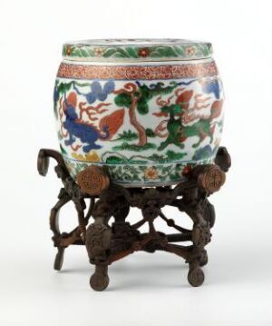  A colorful ceramic jar with floral and animal motifs on a hand-carved dark brown wooden stand. The jar has a green banded top and bottom with a white background and is adorned with blue, orange, and green designs.