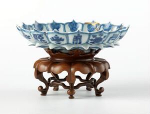  An elegant blue and white porcelain bowl with a scalloped edge and detailed landscape scenes inside, resting on a decorative, carved wooden stand with a reddish-brown finish.