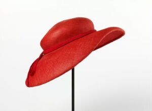  A sophisticated red hat with a smooth, wide brim that curves up on one side, displayed against a light grey background on a thin black stand.