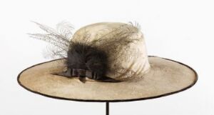  A beige ladies' hat with a round crown and slightly flared brim adorned with dramatic dark gray and black feathers, displayed on a circular stand with a plain, light background.