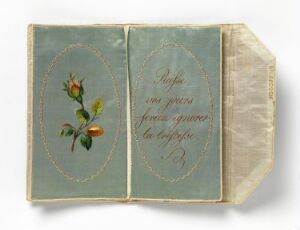  An open vintage book or album with a left page featuring an embroidered red rose with green leaves on a pale blue background and the right page displaying elegant cursive script in silver thread, bordered with cream lace.