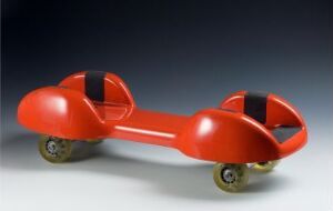  "Multiroller" by Labyrinth Development, a bright red, wheeled plastic design object resembling an elongated dumbbell with concave sections and black straps, on a gray gradient background.