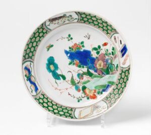  A decorative ceramic plate with a blue central floral motif surrounded by colorful flowers, ringed by a green scalloped border, displayed on a plate stand against a plain backdrop.