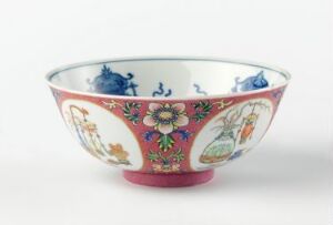  A decorative bowl with a red exterior featuring gold-outlined paneled motifs and floral patterns in blue, green, yellow, and pink. The interior is white with blue decorative elements. The bowl is on a white surface against a light grey background. Artist name and title are unknown.