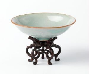  A delicate Asian bowl with a seafoam green to celadon gradient glaze, resting on an ornate dark wooden stand with twisted leg design, against a neutral background. Artist name and title unknown.