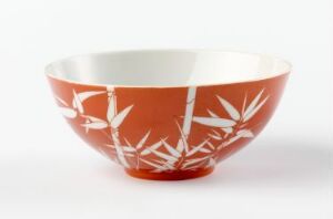  An elegantly shaped bowl with a tapered base and wide opening, coated on the outside in a warm terracotta orange with stylized white bamboo leaf designs, set against a light grey background. The inside of the bowl is glossy white.