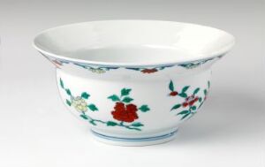  A porcelain bowl with a flared rim, featuring a hand-painted floral design with red flowers, blue flowers, and green leaves against a white background. A thin blue line borders the rim of the bowl, set against a neutral backdrop.