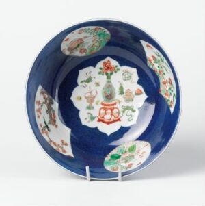  A round ceramic plate with a dark blue outer rim and a central design featuring a white medallion surrounded by fan-shaped panels and smaller oval insets, all adorned with colorful floral patterns in red, green, and blue on a white background. Artist name and title are unknown.