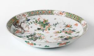  An antique ceramic plate with a detailed, hand-painted floral and avian design in greens, oranges, pinks, and blues on a white background, with a decorative border along the rim featuring green, red, and gold patterns.