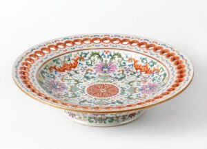  An ornate oval porcelain bowl with scalloped edges, featuring intricate orange, green, blue, and purple floral patterns on a white background, and a central circular terracotta-colored medallion. The bowl is displayed against a stark white backdrop. Artist name and title are unknown.