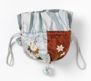  A folded multi-colored fabric face mask with pale sky blue, steel blue, and rusty brown panels, decorated with a white floral pattern on the brown section, white drawstrings, and fluffy white pom-poms on a white background.