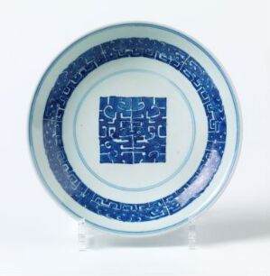  A blue and white porcelain plate with intricate cobalt blue patterns on a white background, standing on a clear stand against a neutral backdrop. The rim of the plate is decorated with a repeating pattern of interconnected geometric shapes and stylized motifs, while the center features a focused grid-like pattern reminiscent of an emblem or character.