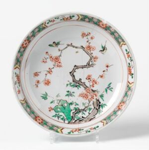  An ornate round plate with a white background, featuring a central cherry blossom branch in pink and orange tones, edged with sections of green foliage and blue and green birds. The plate is intricately detailed and possibly antique, exemplifying fine decorative craftsmanship. Artist name and title are unknown.