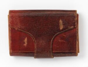  An antique, palm-sized book with a textured reddish-brown leather cover, featuring a curved indentation near the bottom, suggesting frequent use and age. The book is closed and shows signs of wear, with no visible text on the cover.