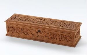  An elaborately carved wooden box with a hinged lid, intricate floral patterns, and a keyhole, resting on a light gray surface against a white background. The box's rich medium-brown wood highlights the craftsmanship of the design.