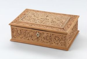  An intricately carved light brown wooden box with a flat hinged lid, featuring detailed floral patterns engraved on all visible surfaces, including a lock keyhole at the front, displayed against a neutral background.