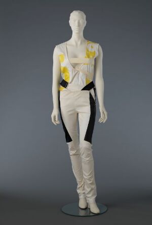  A mannequin dressed in an avant-garde costume by Helmut Lang, with a light base and accents in pale yellow and black, featuring a geometric pattern, a rubber elastic belt with a metal buckle, and pants that extend over the feet.