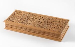  An intricately carved wooden box with a central motif of leaves and florals, surrounded by ornate scrollwork and detailed edges, set against a plain background.