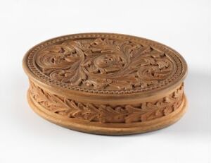 
 A circular wooden box with elaborate floral and scrollwork carvings on the lid and sides, showcasing a light brown color and detailed craftsmanship.