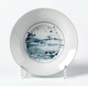  A white porcelain plate with a blue landscape painting in the center, supported by a clear stand against a neutral gray background. The design features shades of blue with abstract elements suggesting a natural scenery. Artist name and title are unknown.