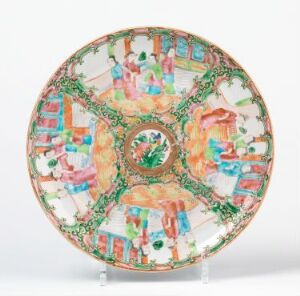  An ornate ceramic plate with an intricate design featuring a central medallion, eight detailed panels with figures and scenes surrounded by green vine-like latticework against a white background, displayed on a metal stand. Artist name and title are unknown.