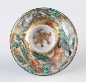  A porcelain bowl with a broad, slightly raised edge, featuring a detailed pattern with elements of nature in gold, green, blue, red, and brown hues, against a light neutral background.