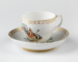  An ornate white porcelain teacup with a matching saucer, featuring a gold rim and a painted scene of a horseback rider with hounds on one side. The gold and colorful illustration contrast elegantly against the white background.