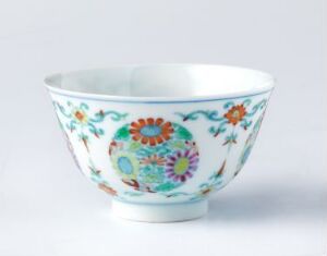  An ornate porcelain bowl with a white background and a colorful, intricate design featuring floral patterns and foliage in shades of blue, orange, red, yellow, and green. The bowl has a broad, elegant body tapering to a small base. Artist name and title are unknown.