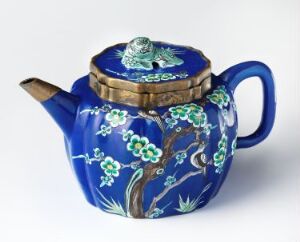  An ornate cobalt blue ceramic teapot decorated with white flowers and green leaves on brown branches, featuring a three-dimensional green frog on the lid.