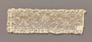  A rectangular piece of antique cream lace with a floral design on a grey background.