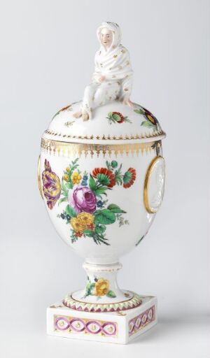  A decorative white porcelain urn with a seated dog figure on the lid, featuring vibrant floral patterns in purple, red, yellow, pink, and green, with gold accents and a square base adorned with pink and gold details, against a light grey background. Artist name and title unknown.