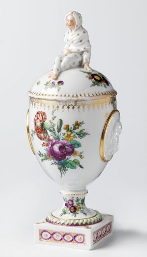  A small ornate porcelain urn adorned with a bird figure on top and painted with purple fruit, green leaves, and classical gold-trimmed motifs over a white background. There is no artist's name or title provided.