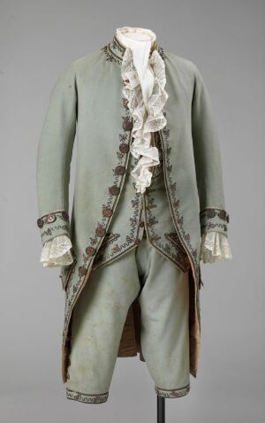  A historical men's suit from the 18th or early 19th century, displayed on a mannequin, featuring a muted pistachio green coat with forest green and gold buttons and embroidered cuffs, paired with matching breeches and a white ruffled jabot, set against a neutral gray background.