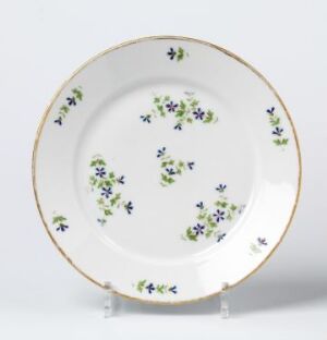  A white ceramic plate with a gold rim featuring a scattered pattern of blue flowers and green leaves, displayed on a transparent stand against a neutral background. Artist name and title are unknown.