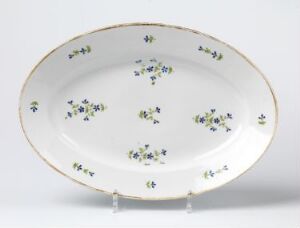  An oval porcelain plate with a white base and a gold rim, decorated with small clusters of blue, yellow, and green flowers, displayed on a stand against a neutral background.