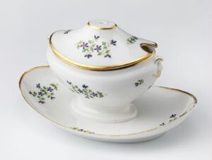  A white porcelain sugar bowl with a lid and gold trim resting on a matching saucer, both decorated with small blue and green floral patterns, set against a plain background. Artist name and title are unknown.