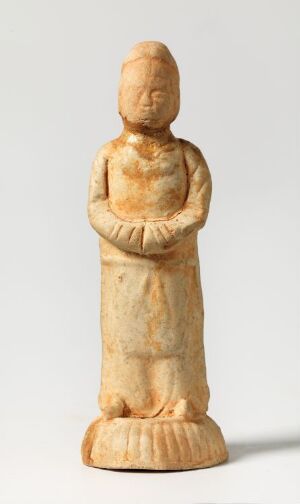  A small, simplistic figurine carved from light beige material depicting a standing human figure with folded arms and minimal facial features, set against a white background. Artist name and title are unknown.