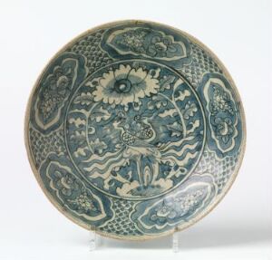  An antique blue and white circular ceramic plate with intricate floral and foliate patterns, displayed on a stand against a neutral backdrop. Artist name and title are unknown.