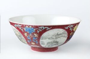  A porcelain bowl with a deep red exterior featuring oval monochrome landscape reserves and colorful floral motifs, contrasted by a pure white interior, set against a light-grey background.