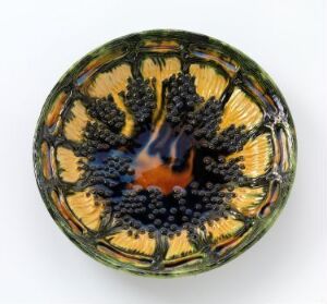  Top-down view of a decorative bowl with a symmetrical pattern featuring a burnt orange center, surrounded by clusters of black dots and a gradient background from golden yellow to olive green. Artist name and title unknown.