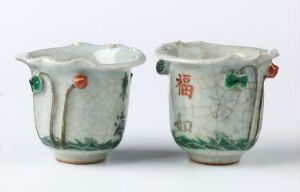  Two small, antique-looking ceramic cups with celadon glaze, crackled texture, and animal head-shaped handles in green, orange, and red. The rightmost cup features a red character or symbol on its side.