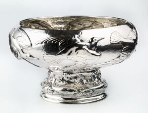  A polished silver bowl with embossed fish design, exhibiting a gleaming metallic surface with light reflecting and creating highlights and shadows, displayed against a neutral background.