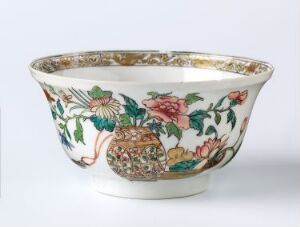 
 An ornate porcelain bowl with a wide rim, detailed with a floral design including a large pink peony, smaller flowers and greenery on a white background, all accented with a golden decorative border around the rim. Artist name and title are unknown.