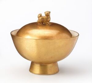  A golden, polished metal bowl with a matching lid that has a small animal figure as a handle on top, reflecting a simple yet elegant design.