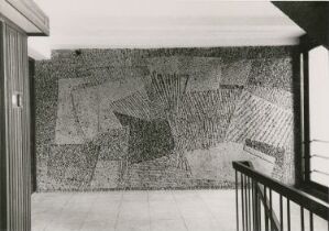  A black and white photograph of a modern architectural interior featuring a large, textural mosaic wall in a pattern of contrasting dark and light elements. To the left is a partially open door, and the right side shows a railing, possibly next to stairs. The flooring is tiled and reflective, suggesting a polished stone material. The ambient lighting creates a calm atmosphere, highlighting the clean, minimalist design.