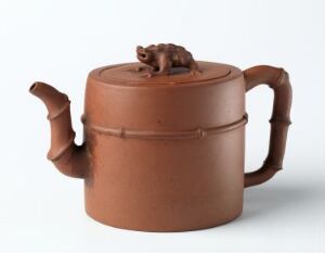  A traditional round-bodied terra cotta teapot with a sculpted frog on the lid and two spouts. It features a warm brown color, two handles, and an encircling indentation in the middle, set against an off-white background.