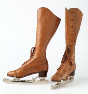  Vintage caramel-brown leather ice skates with elongated boots and steel blades, set against an off-white background. The skates feature decorative leather loops for lacing and are mounted on a wooden sole attached to the metal blade.