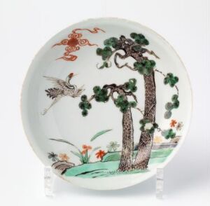  A decorative plate featuring a painted scene with a brown tree with green foliage, a bird in flight, and clusters of orange and red flowers, set against a glossy white background. Artist name and title are unknown.